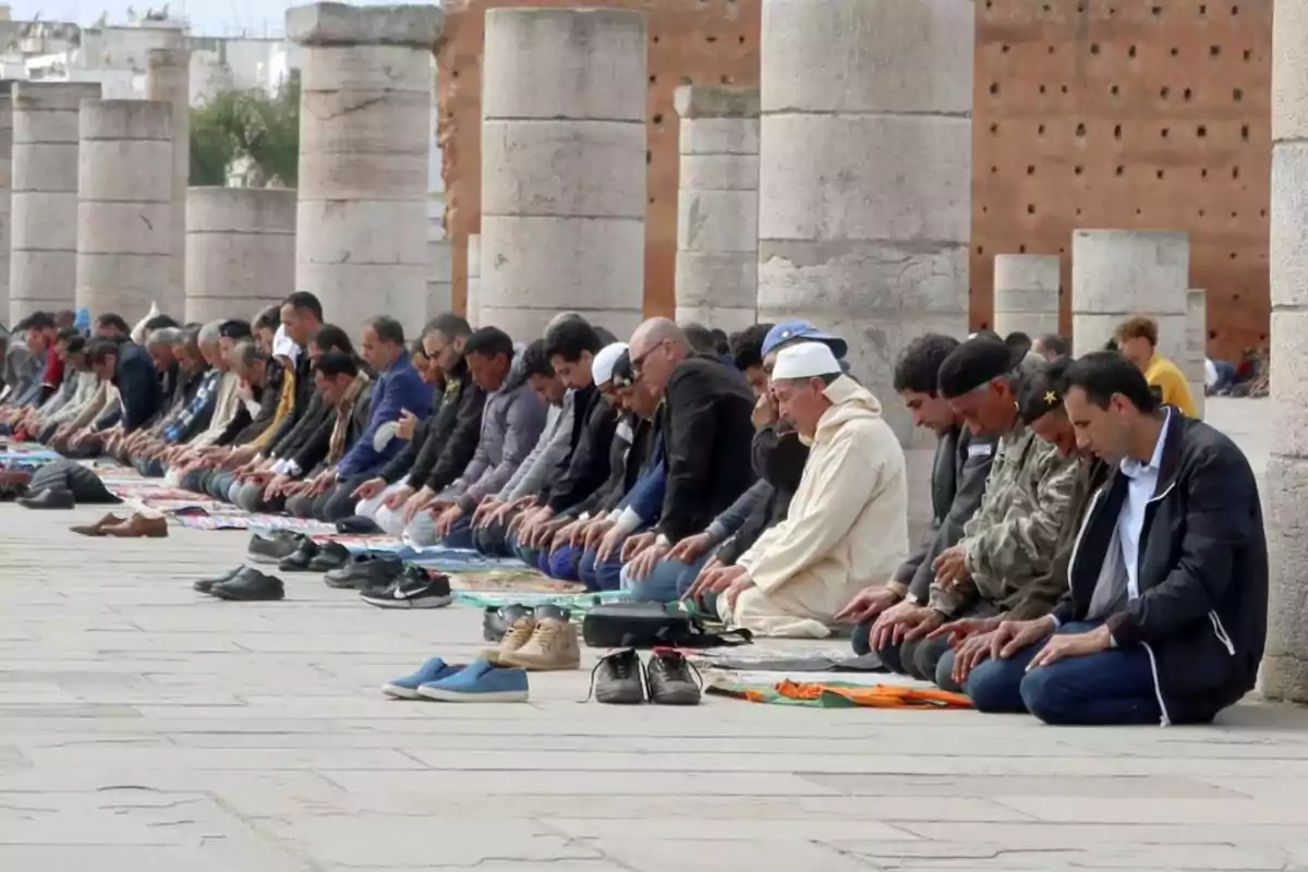 Un grupo de personas rezando al aire libre en una fila, con columnas de piedra al fondo.