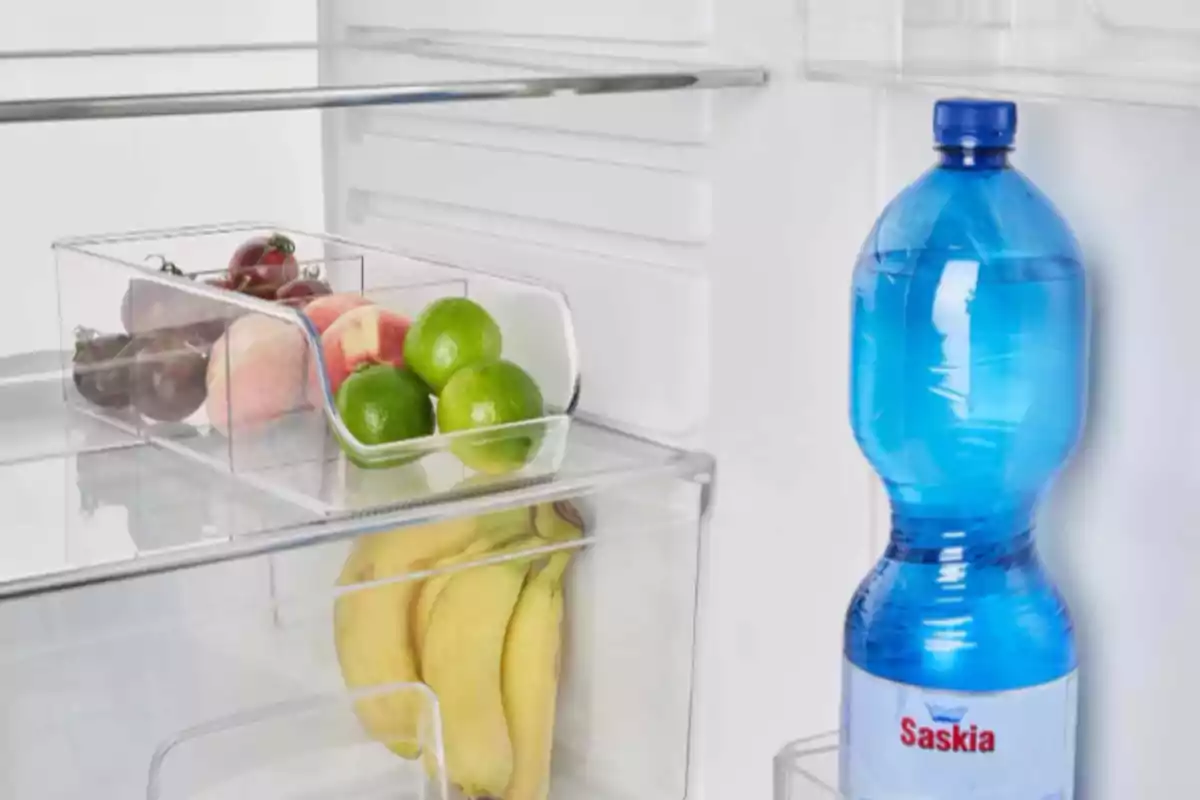 Una nevera abierta muestra una botella de agua azul en la puerta y un cajón con frutas como limones, duraznos y tomates, mientras que en otro compartimento se ven plátanos. Una nevera abierta muestra una botella de agua azul en la puerta y un cajón con frutas como limones, duraznos y tomates, mientras que en otro compartimento se ven plátanos.