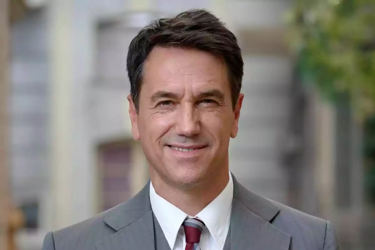 Oriol Tarrasón sonriendo con traje gris y corbata roja en un entorno urbano en Amar es para siempre. Oriol Tarrasón sonriendo con traje gris y corbata roja en un entorno urbano en Amar es para siempre.