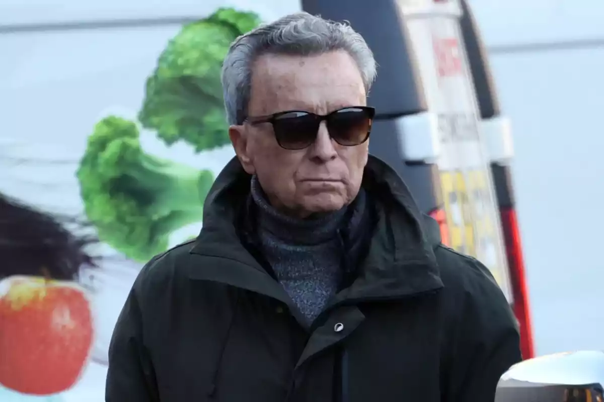 Ortega Cano con gafas de sol y abrigo oscuro camina al aire libre con un fondo de imágenes de frutas y verduras. Ortega Cano con gafas de sol y abrigo oscuro camina al aire libre con un fondo de imágenes de frutas y verduras.
