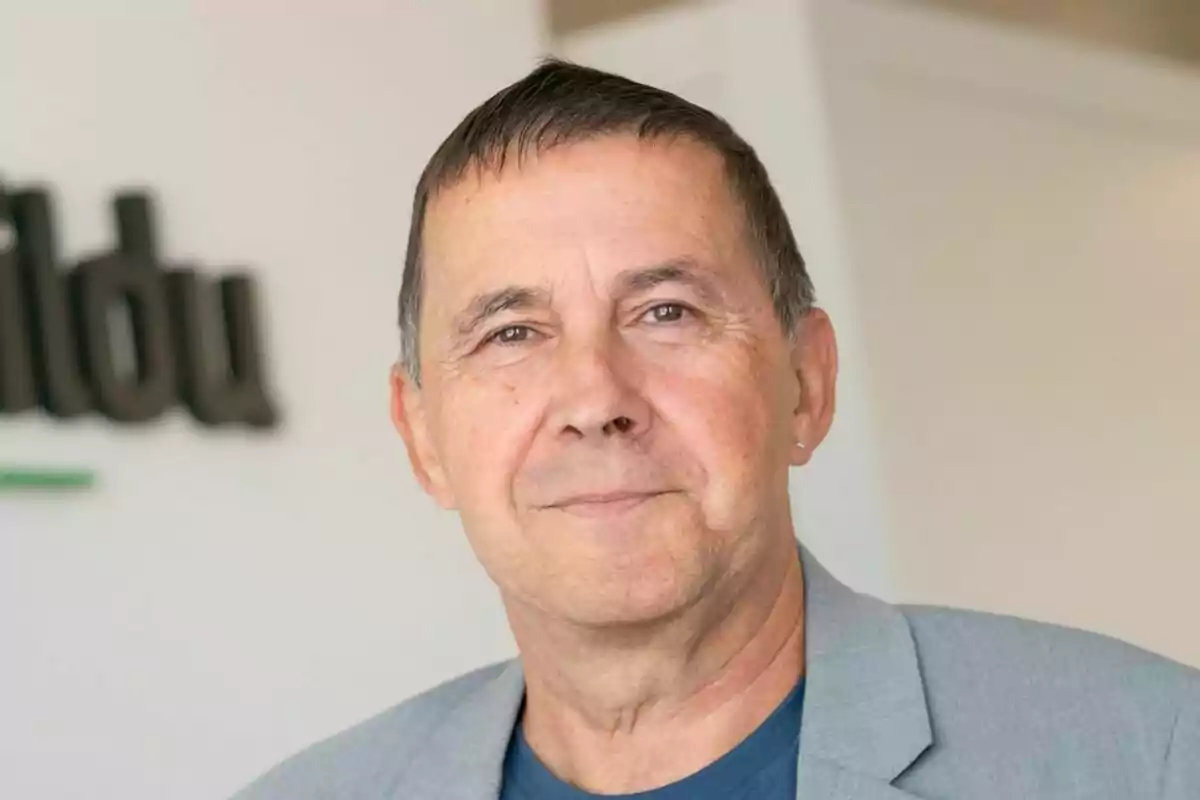 Un hombre de mediana edad con cabello corto y una chaqueta gris sonríe ligeramente frente a un fondo claro con un letrero borroso.