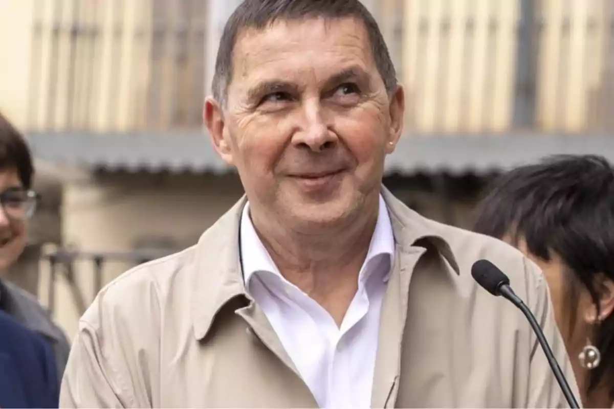 A man in a beige jacket and white jersey smiles while standing in front of a microphone with people in the background.