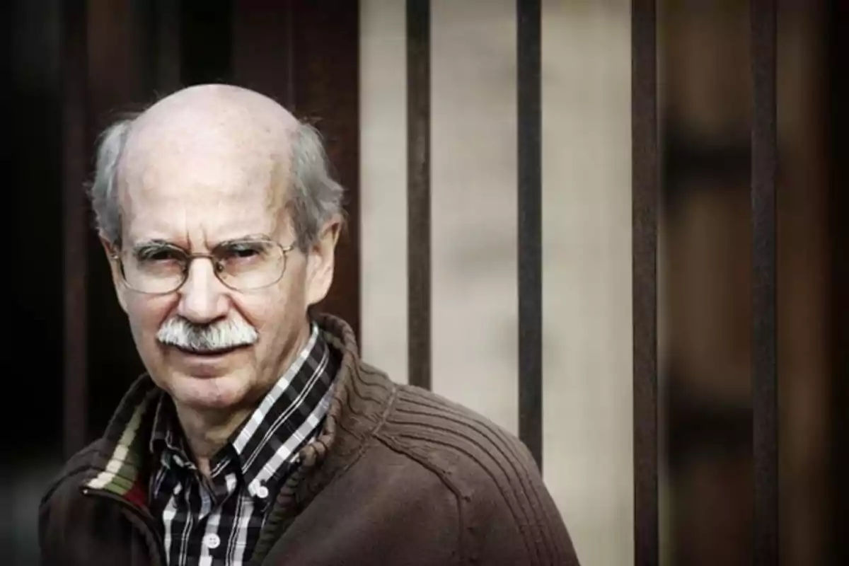 Un hombre mayor con gafas y bigote viste una camisa a cuadros y un suéter marrón mientras está de pie frente a una estructura de barras metálicas.