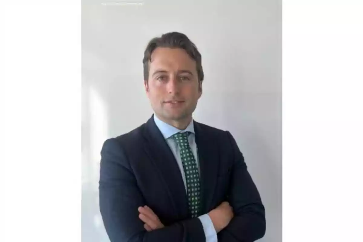 Hombre joven de cabello castaño con traje oscuro y corbata verde posando con los brazos cruzados frente a una pared blanca