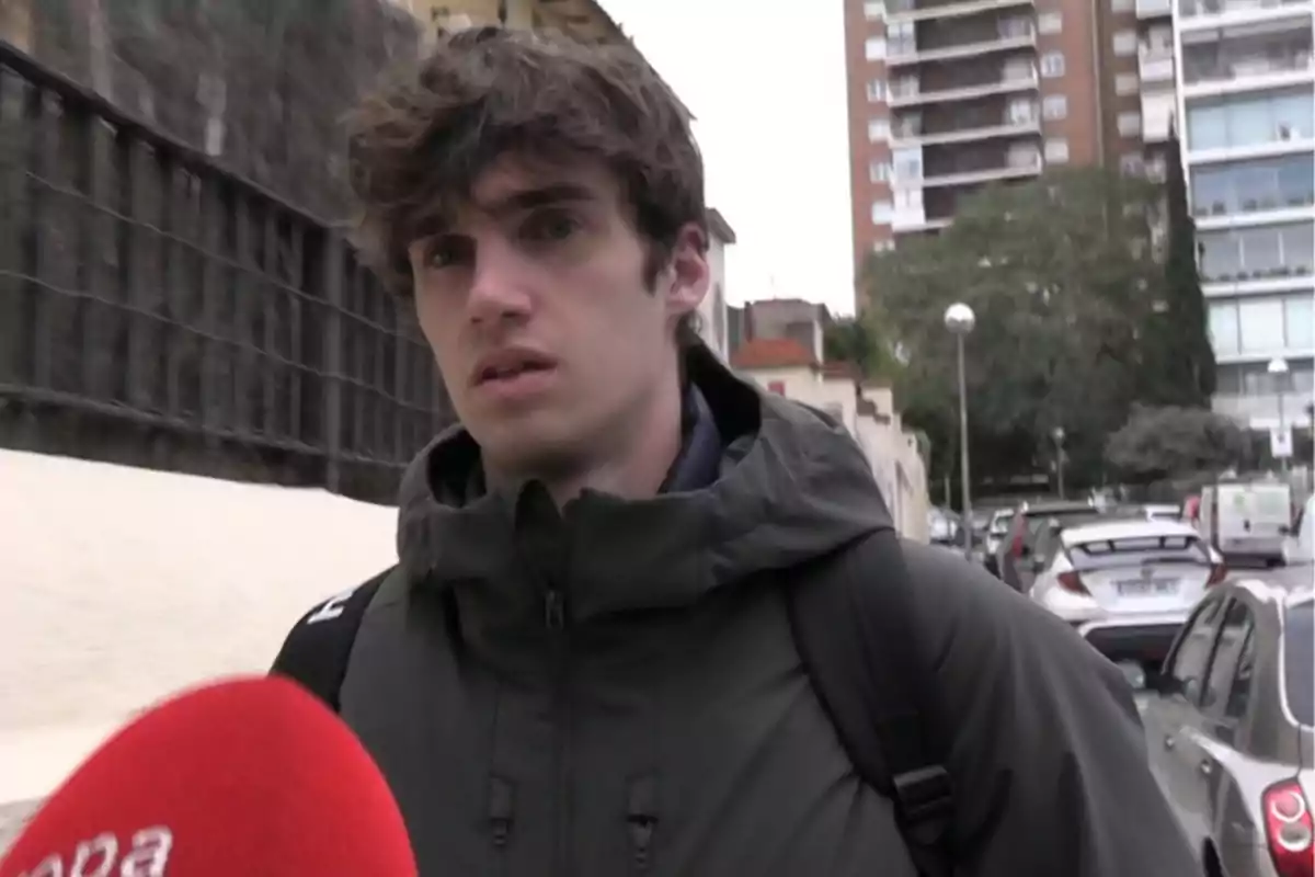 Pablo Urdangarin wearing a dark jacket and backpack is interviewed on an urban street with buildings and cars in the background. Pablo Urdangarin wearing a dark jacket and backpack is interviewed on an urban street with buildings and cars in the background.
