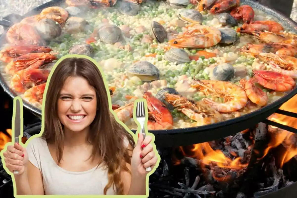 Una mujer sonriente sostiene un tenedor y un cuchillo frente a una gran paella de mariscos cocinándose sobre fuego.