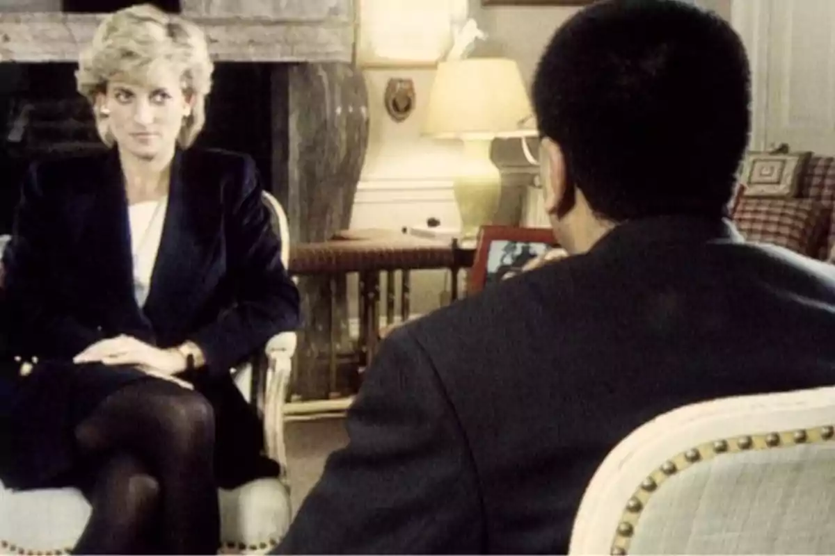 Lady Di sentada en una silla, vestida con un traje oscuro, parece estar en una entrevista con un hombre de espaldas, en una habitación con una lámpara y una chimenea al fondo. Lady Di sentada en una silla, vestida con un traje oscuro, parece estar en una entrevista con un hombre de espaldas, en una habitación con una lámpara y una chimenea al fondo.