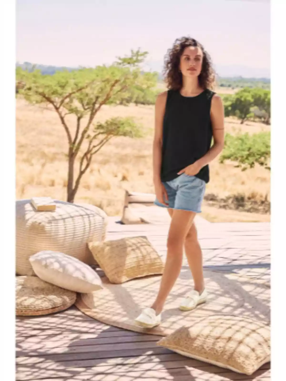 Mujer de cabello rizado con camiseta negra y shorts de mezclilla caminando sobre una terraza de madera decorada con cojines y pufs en un entorno natural y soleado Mujer de cabello rizado con camiseta negra y shorts de mezclilla caminando sobre una terraza de madera decorada con cojines y pufs en un entorno natural y soleado