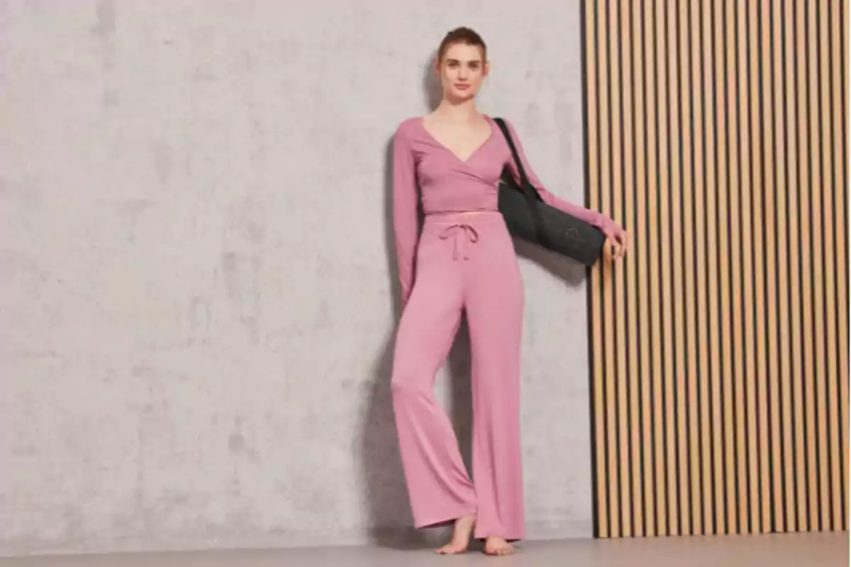 Una persona con un conjunto rosa y un bolso negro posa descalza junto a una pared de concreto y paneles de madera. Una persona con un conjunto rosa y un bolso negro posa descalza junto a una pared de concreto y paneles de madera.