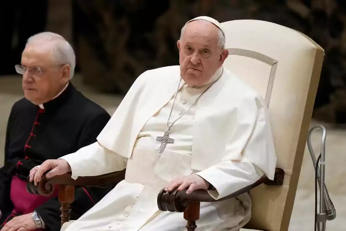 Un líder religioso vestido de blanco está sentado en una silla junto a otro clérigo.