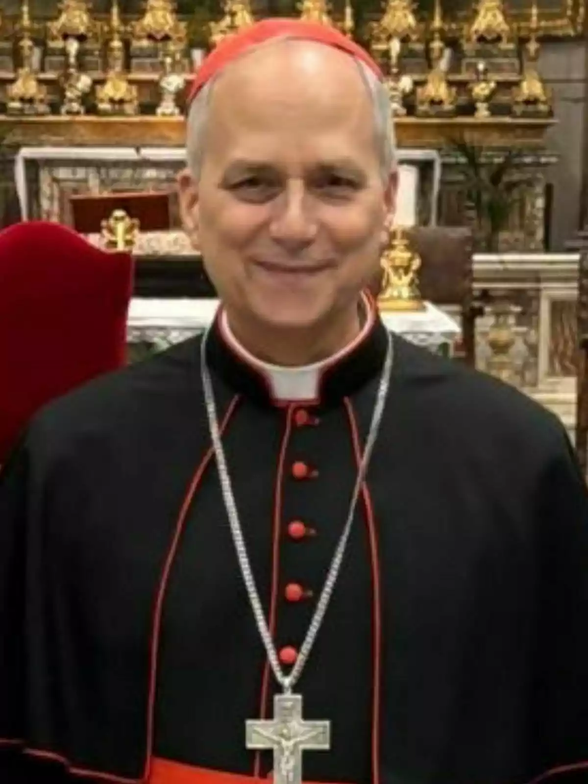 Pope Leo XIV dressed in black and red ecclesiastical attire with a cross hanging from his neck smiles in front of an altar decorated with golden ornaments. Pope Leo XIV dressed in black and red ecclesiastical attire with a cross hanging from his neck smiles in front of an altar decorated with golden ornaments.