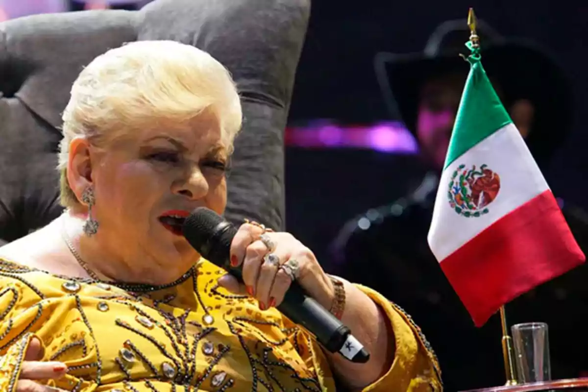 A person with blonde hair sings into a microphone while sitting in an armchair next to a Mexican flag.