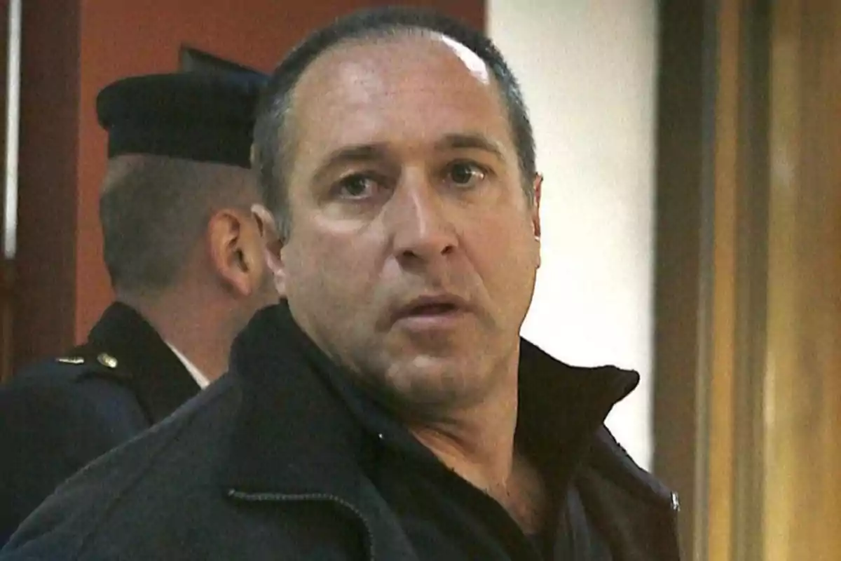 Hombre de cabello corto y expresión seria en un entorno interior con otra persona uniformada al fondo
