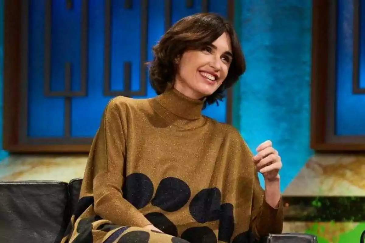 Paz Vega con cabello oscuro y corto sonríe mientras está sentada en un sofá con un fondo azul en La Revuelta.