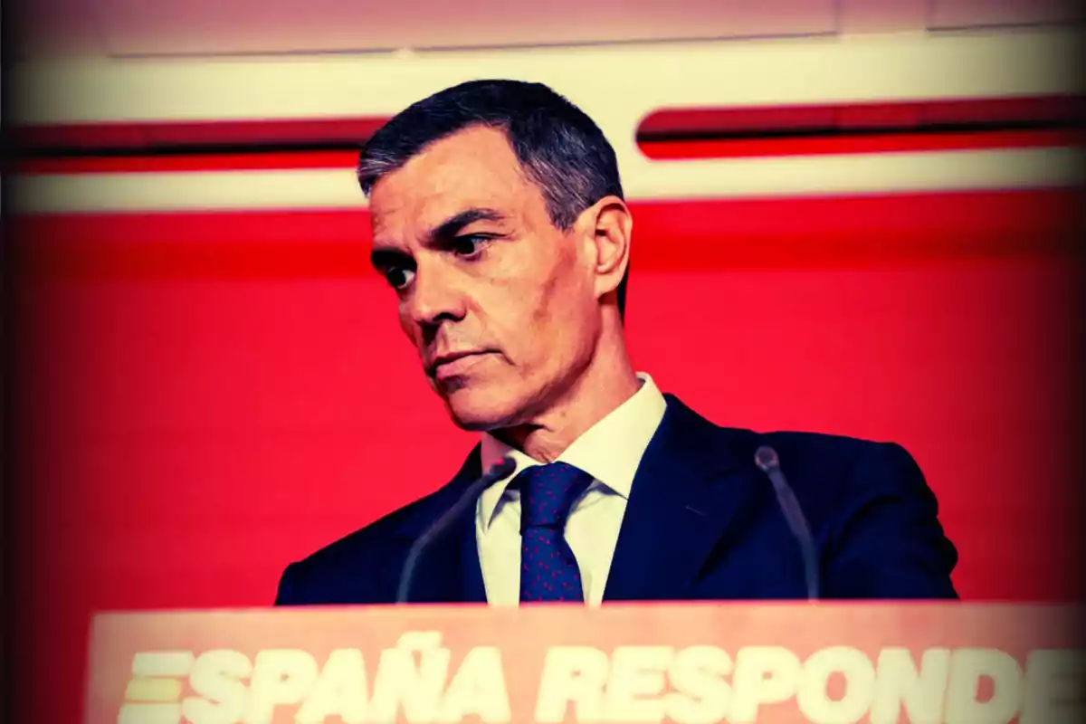 Hombre de traje oscuro y corbata azul con fondo rojo y un cartel que dice España responde Hombre de traje oscuro y corbata azul con fondo rojo y un cartel que dice España responde