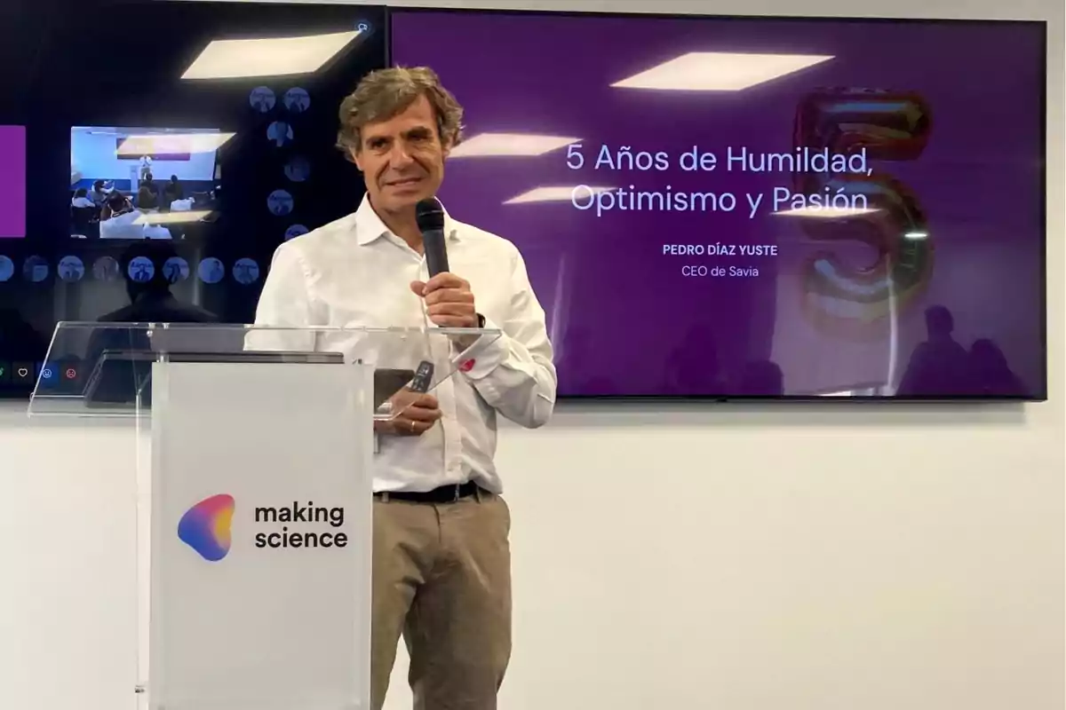 Un hombre con camisa blanca y pantalones beige sostiene un micrófono mientras se encuentra detrás de un podio con el logotipo de "making science". Detrás de él, una pantalla muestra el texto "5 Años de Humildad, Optimismo y Pasión" junto con el nombre "Pedro Díaz Yuste, CEO de Savia".