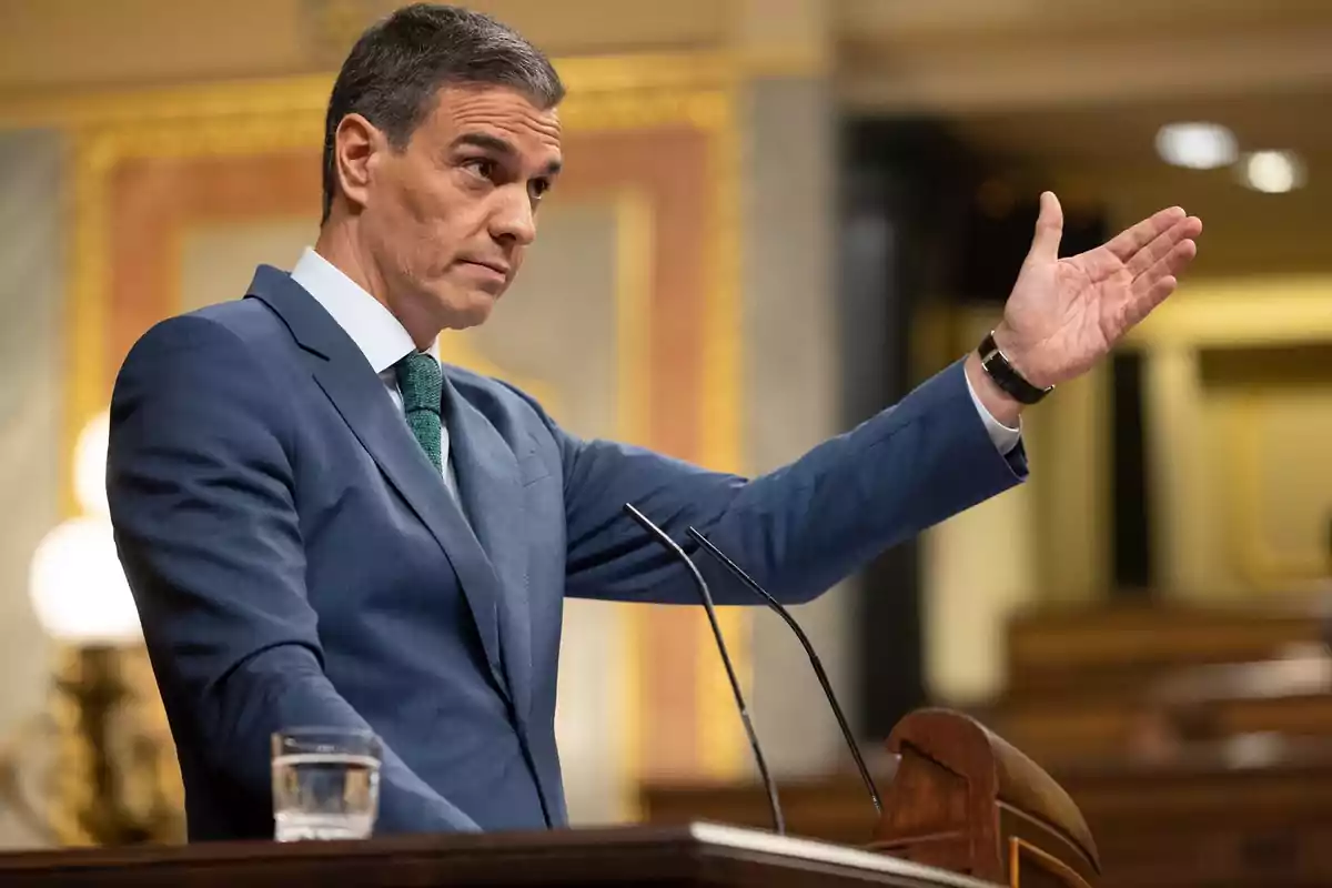 Hombre en traje azul hablando en un podio con un micrófono y un vaso de agua.