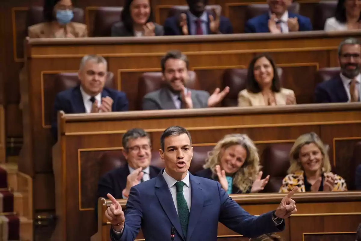 Un hombre en un traje oscuro está hablando en un podio mientras varias personas sentadas detrás de él aplauden y sonríen en un entorno formal. Un hombre en un traje oscuro está hablando en un podio mientras varias personas sentadas detrás de él aplauden y sonríen en un entorno formal.