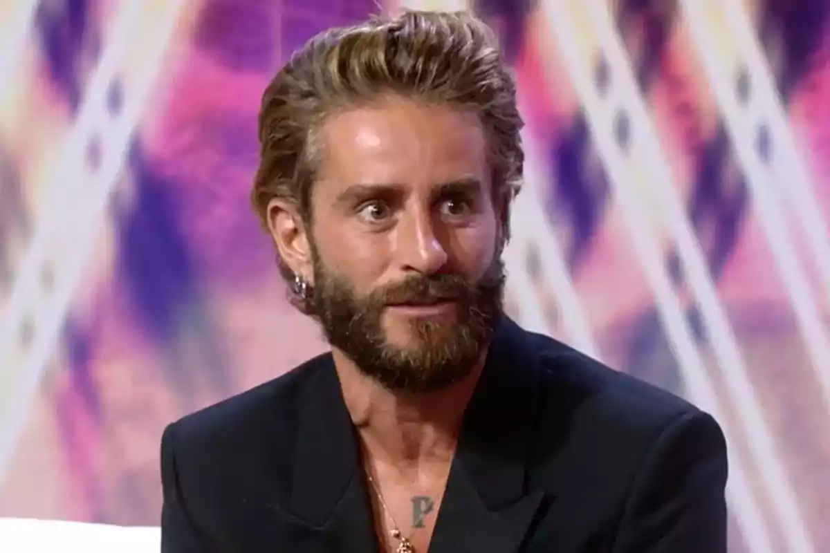 Pelayo Díaz con barba y cabello peinado hacia atrás vistiendo saco oscuro en un set de televisión con fondo colorido Pelayo Díaz con barba y cabello peinado hacia atrás vistiendo saco oscuro en un set de televisión con fondo colorido