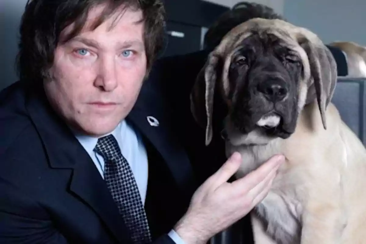 Un hombre de traje oscuro posa junto a un perro grande de color claro, sosteniéndolo con una mano.