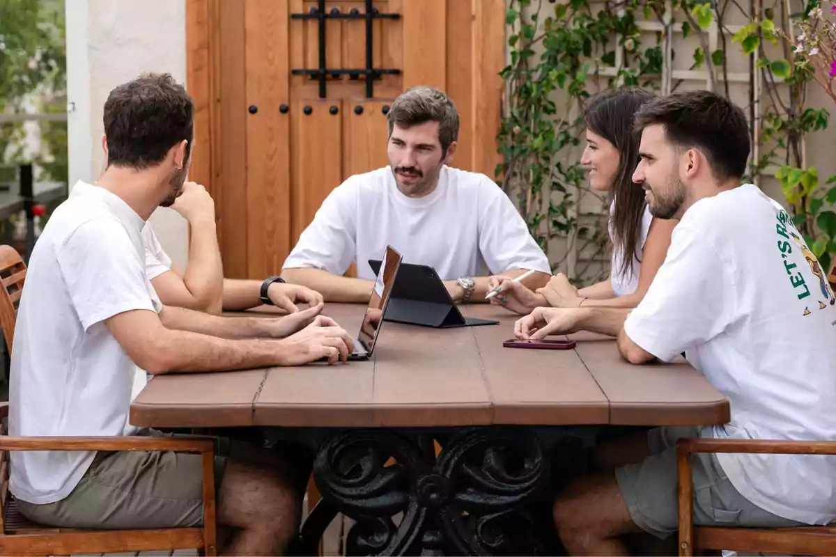 Grupo de personas sentadas alrededor de una mesa al aire libre, trabajando en computadoras portátiles y tabletas.