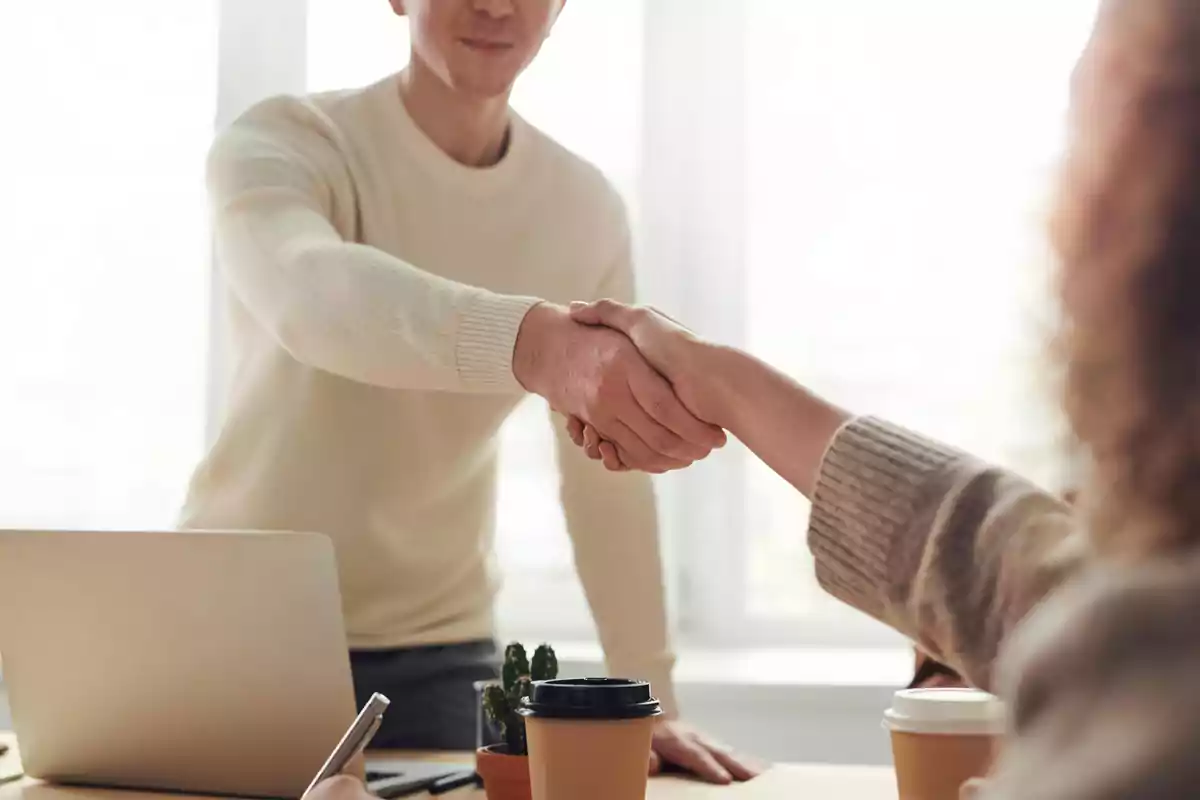 Dos personas dándose la mano en una oficina con una computadora portátil y una taza de café sobre la mesa.