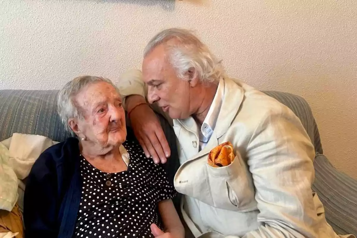 Un hombre de cabello canoso abraza y conversa con una mujer mayor sentados en un sofá.