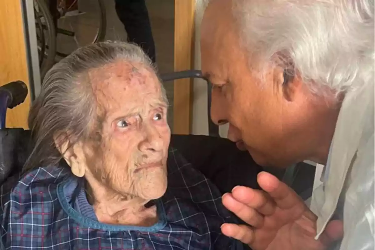 Una persona mayor en silla de ruedas mira a un hombre de cabello canoso que le habla de cerca.