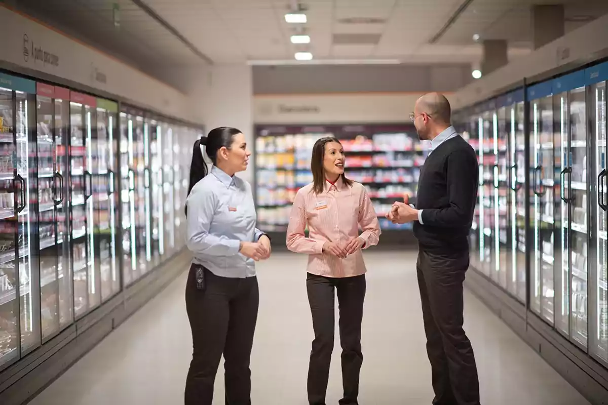 Three people are talking in the frozen foods aisle of a supermarket.