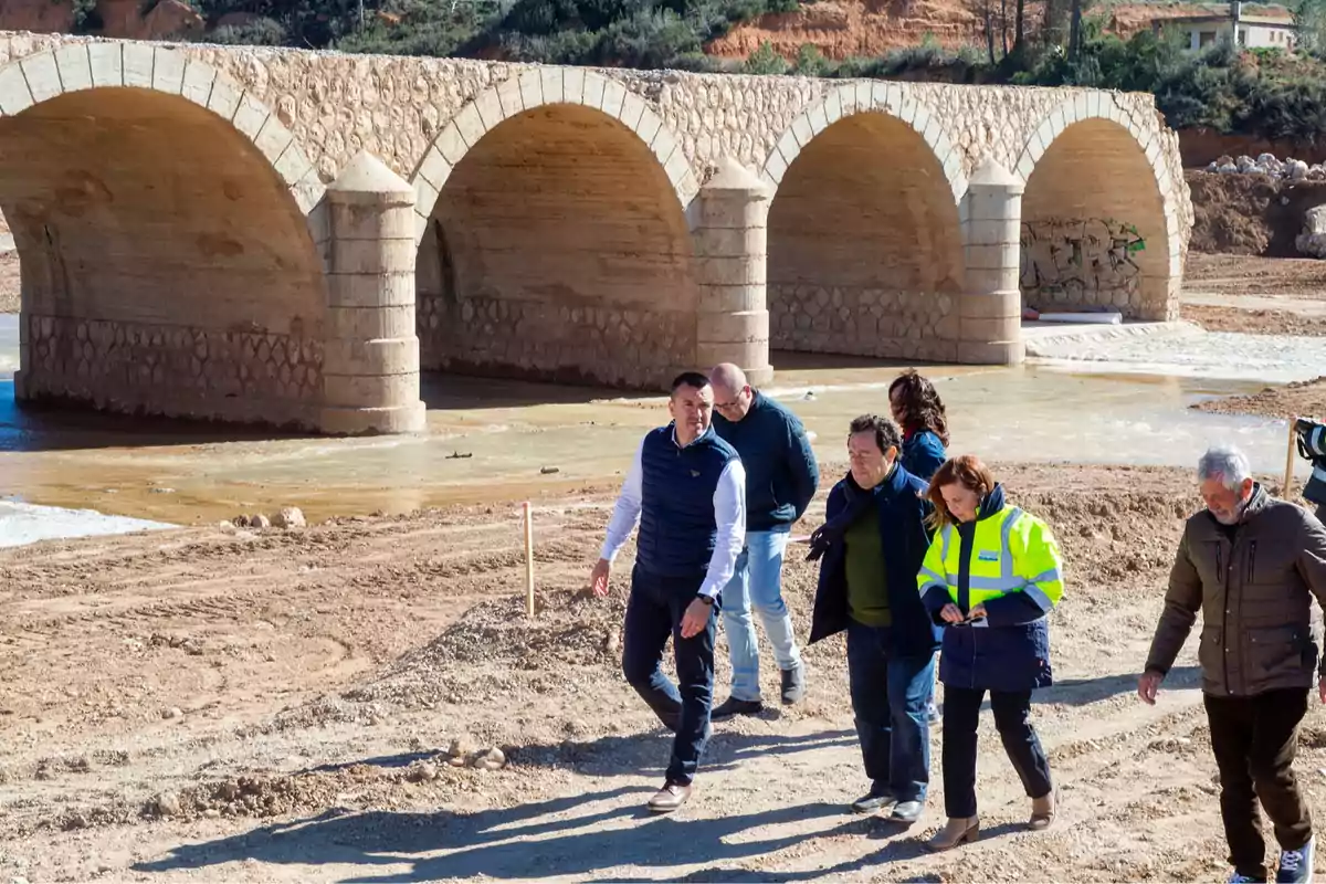 A group of people walks by a stone bridge with several arches over sandy terrain.