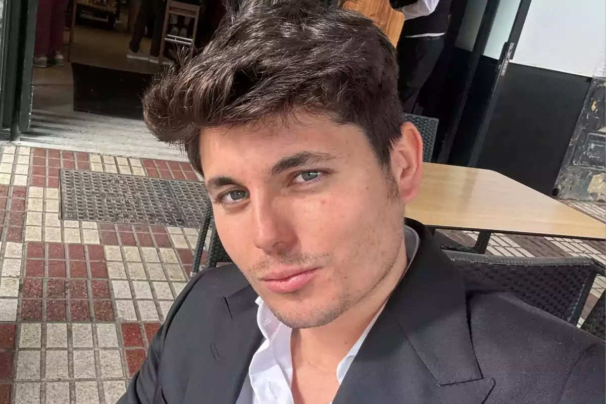 A man with dark hair and a black jacket is sitting on an outdoor terrace.