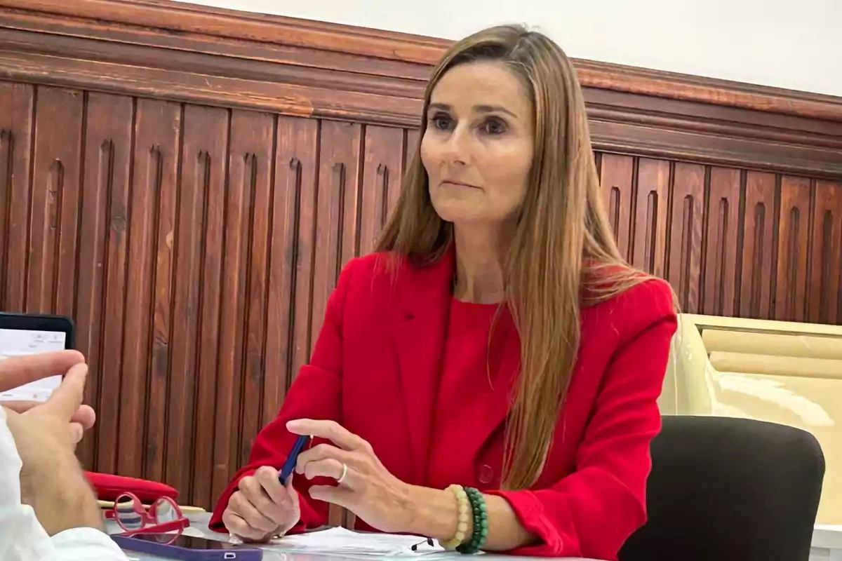 A woman in a red jacket is sitting at a table, holding a pen, in front of a wooden wall.