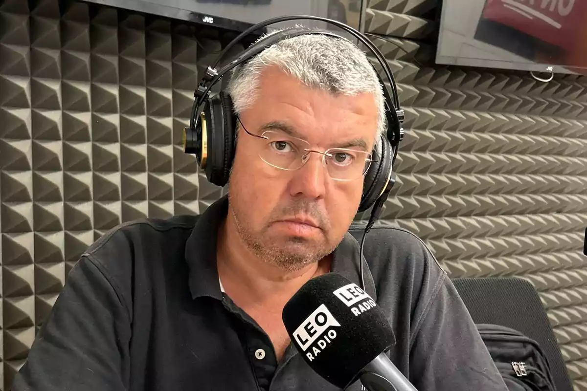 Hombre de cabello canoso y gafas usando audífonos frente a un micrófono en una cabina de radio