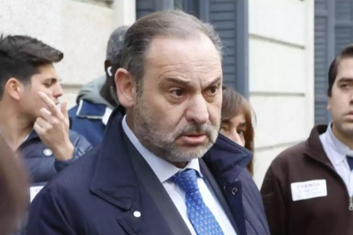 Un hombre con barba y traje azul está rodeado de personas en un entorno urbano.