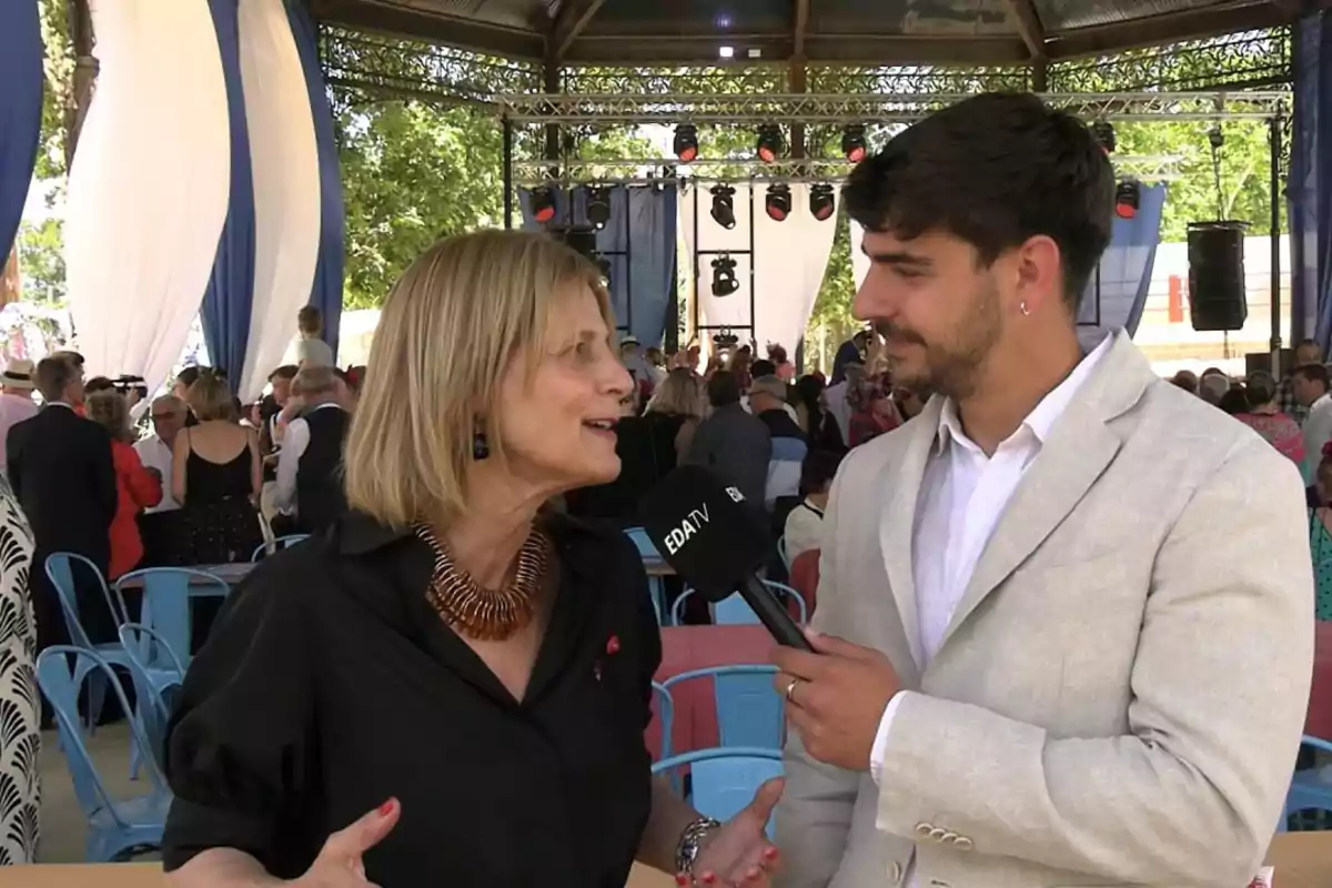 Una mujer es entrevistada por un hombre con micrófono en un evento al aire libre con varias personas y decoraciones en el fondo Una mujer es entrevistada por un hombre con micrófono en un evento al aire libre con varias personas y decoraciones en el fondo