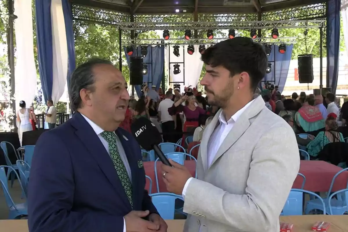 Un reportero entrevista a un hombre con traje azul en un evento al aire libre con gente y decoraciones festivas al fondo Un reportero entrevista a un hombre con traje azul en un evento al aire libre con gente y decoraciones festivas al fondo