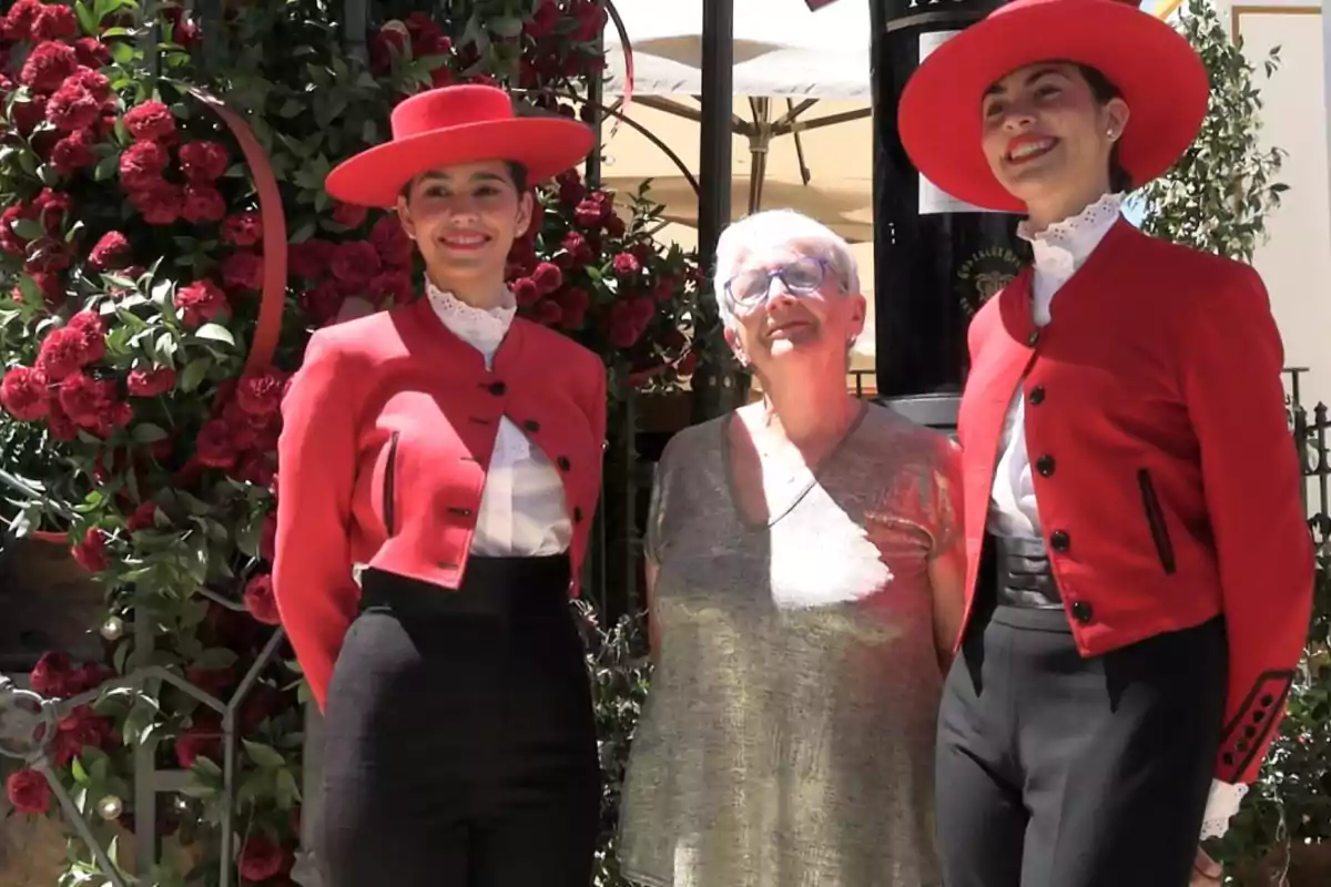 Tres mujeres posan sonrientes al aire libre, dos de ellas llevan trajes tradicionales rojos y sombreros mientras la tercera viste ropa casual y gafas, con un fondo de flores rojas Tres mujeres posan sonrientes al aire libre, dos de ellas llevan trajes tradicionales rojos y sombreros mientras la tercera viste ropa casual y gafas, con un fondo de flores rojas