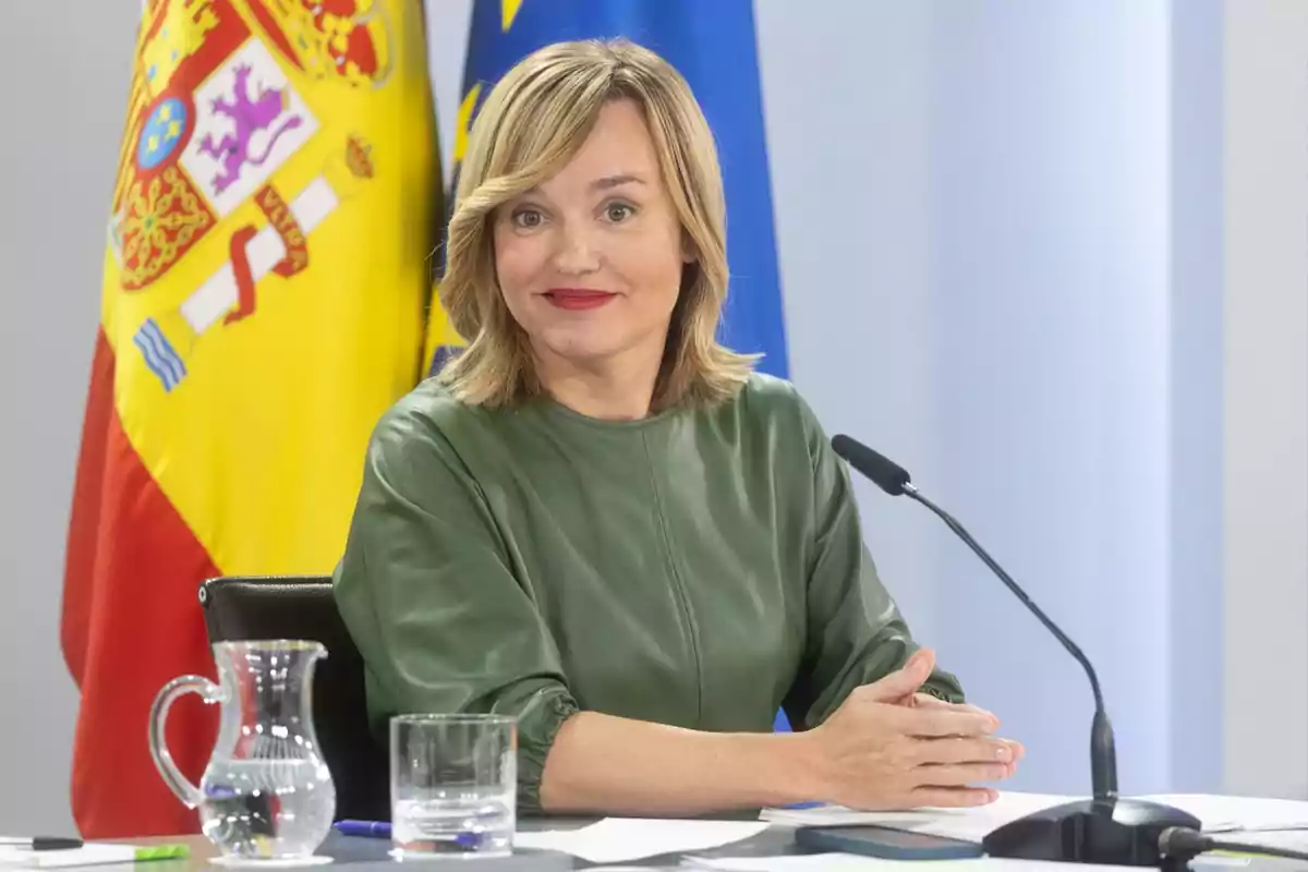 Una mujer con vestido verde sentada frente a un micrófono con banderas de España y la Unión Europea de fondo.