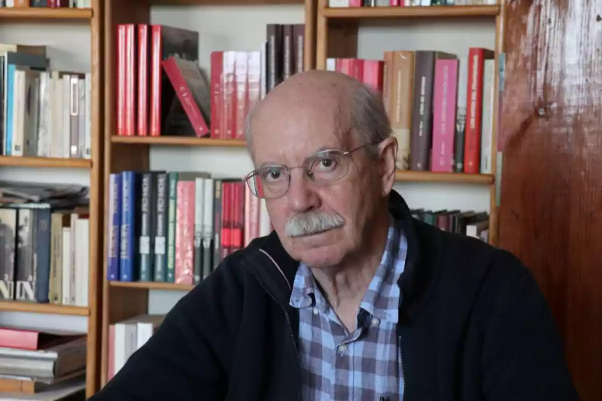 Hombre mayor con gafas y bigote sentado frente a una estantería llena de libros.