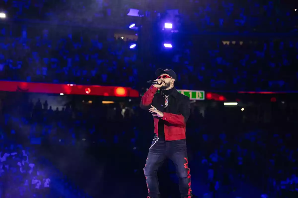 Un cantante con gafas de sol y gorra, vestido con chaqueta roja y negra, actuando en un escenario iluminado con luces moradas frente a una multitud en un estadio.