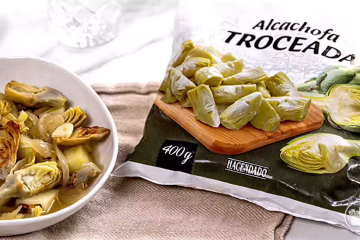 Bolsa de alcachofa troceada Hacendado de 400 gramos junto a un plato con alcachofas cocinadas y rodajas de ajo Bolsa de alcachofa troceada Hacendado de 400 gramos junto a un plato con alcachofas cocinadas y rodajas de ajo