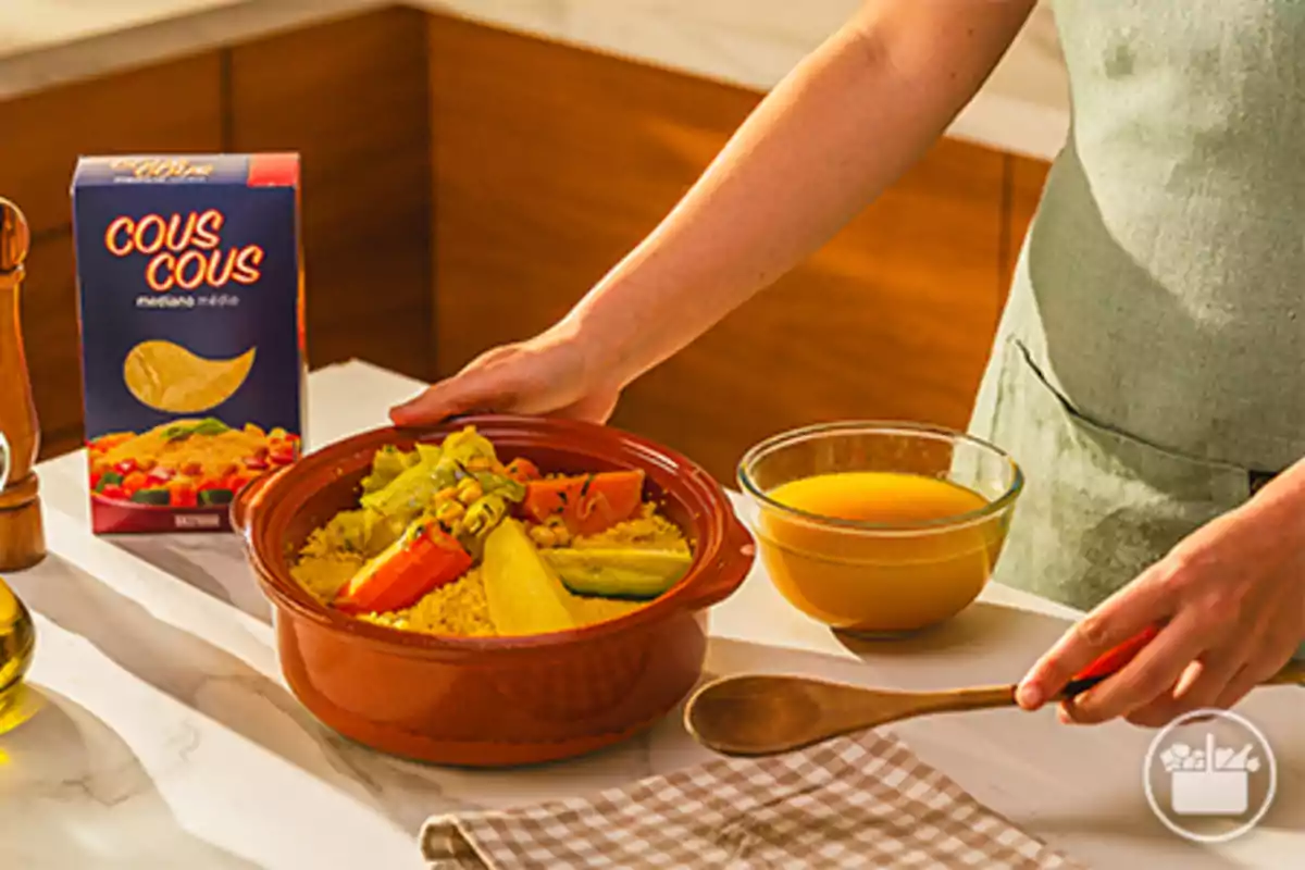 Persona sirviendo cous cous con verduras en un plato de barro junto a un bol de caldo y una caja de cous cous en una cocina Persona sirviendo cous cous con verduras en un plato de barro junto a un bol de caldo y una caja de cous cous en una cocina