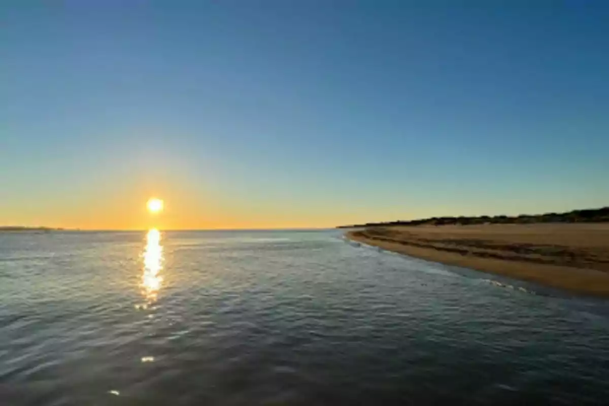 Atardecer en la playa con el sol reflejándose en el agua y un cielo despejado.