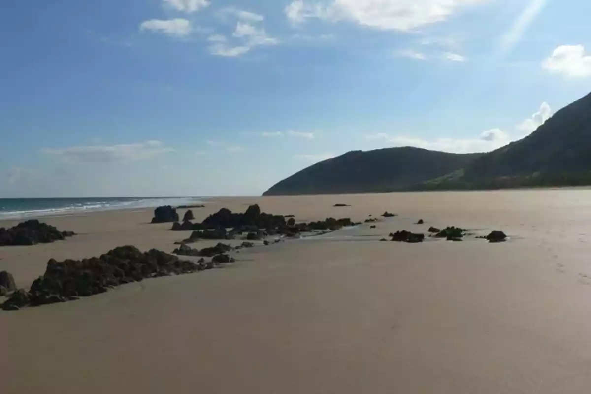 Playa desierta con rocas y montañas al fondo bajo un cielo parcialmente nublado.