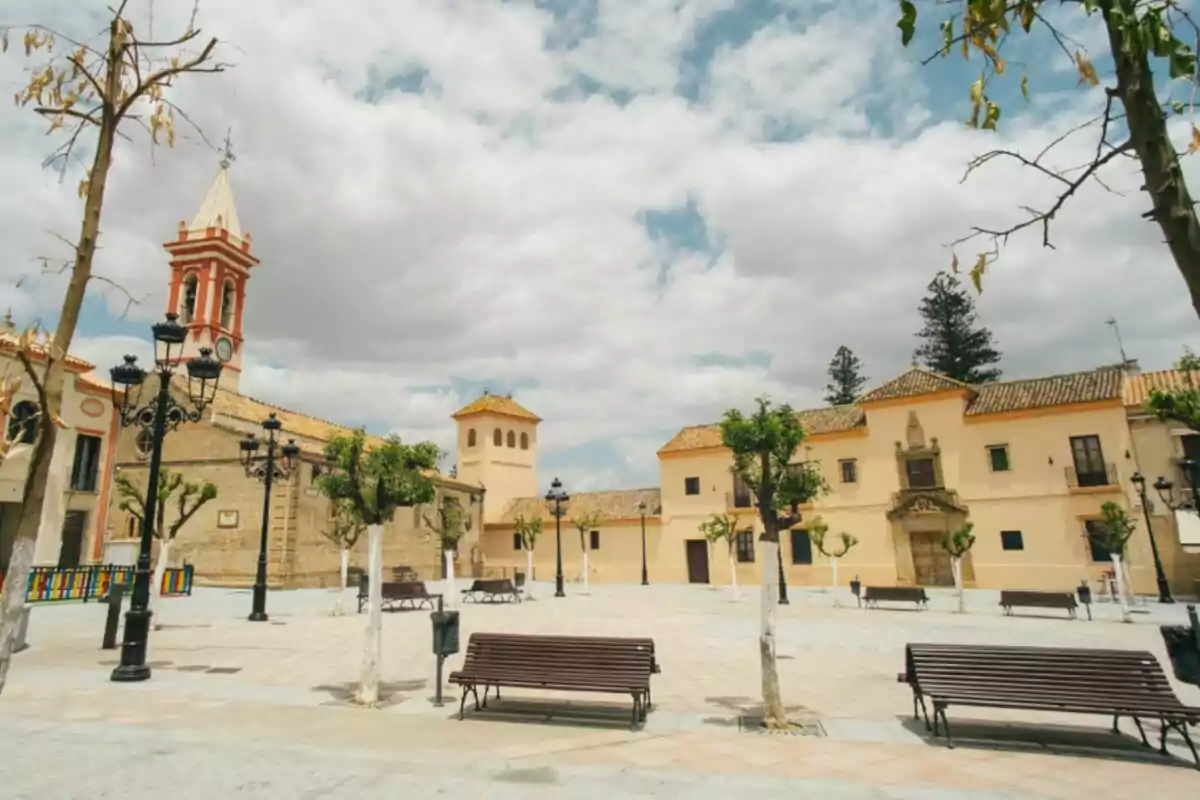 Plaza amplia con bancos, árboles podados, farolas negras y edificios históricos de fondo bajo un cielo parcialmente nublado Plaza amplia con bancos, árboles podados, farolas negras y edificios históricos de fondo bajo un cielo parcialmente nublado