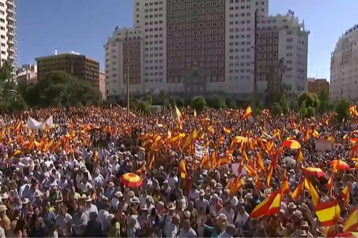 Multitud de personas reunidas en una plaza ondeando banderas de España frente a edificios altos en un día soleado
