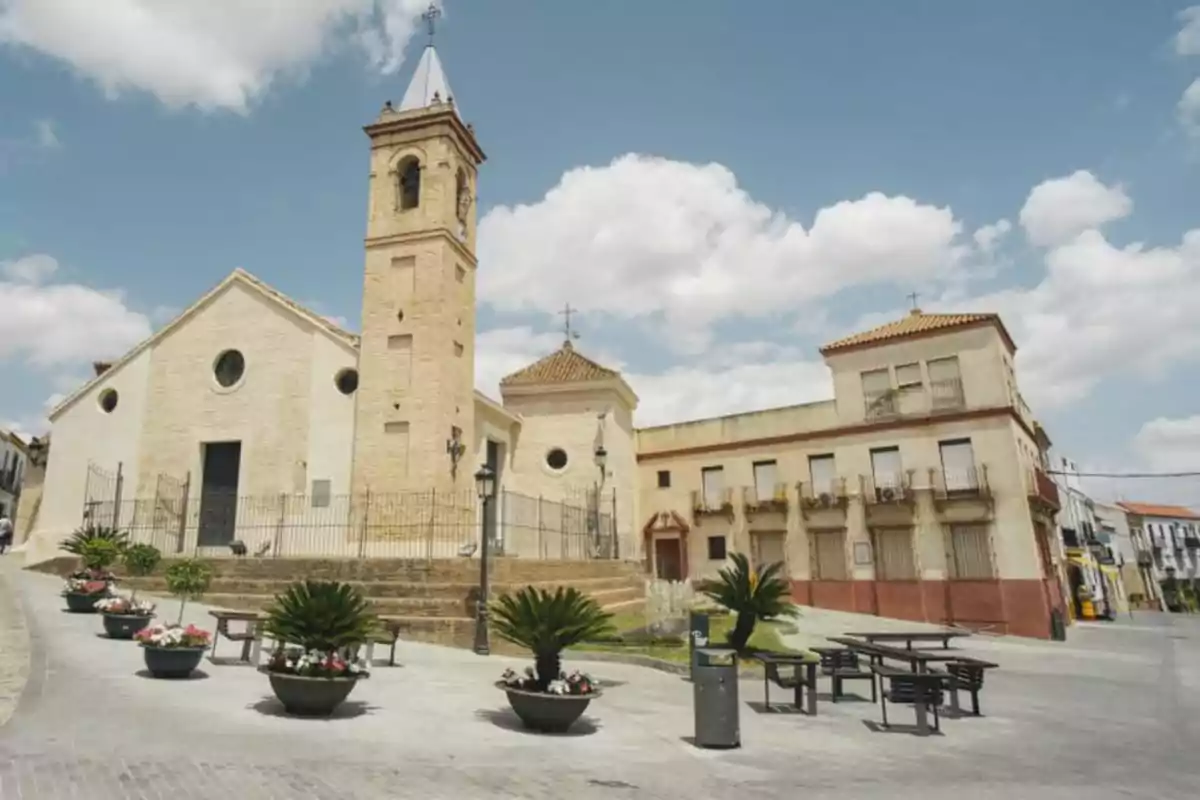 Una iglesia de piedra con una torre y un reloj en una plaza con bancos y macetas bajo un cielo azul con nubes. Una iglesia de piedra con una torre y un reloj en una plaza con bancos y macetas bajo un cielo azul con nubes.