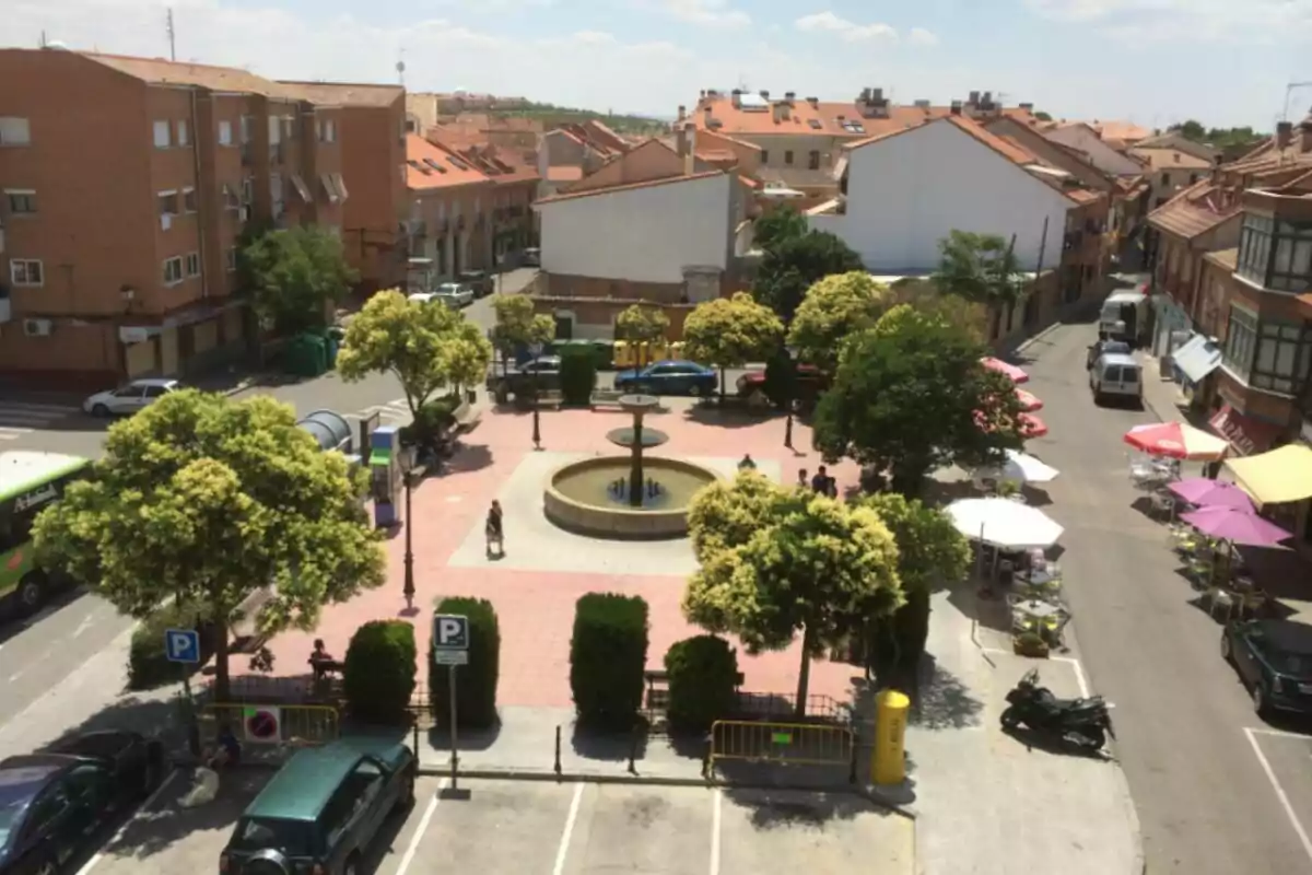 Una plaza con una fuente central rodeada de árboles y bancos, con edificios de ladrillo alrededor y varias mesas con sombrillas en la acera. Una plaza con una fuente central rodeada de árboles y bancos, con edificios de ladrillo alrededor y varias mesas con sombrillas en la acera.