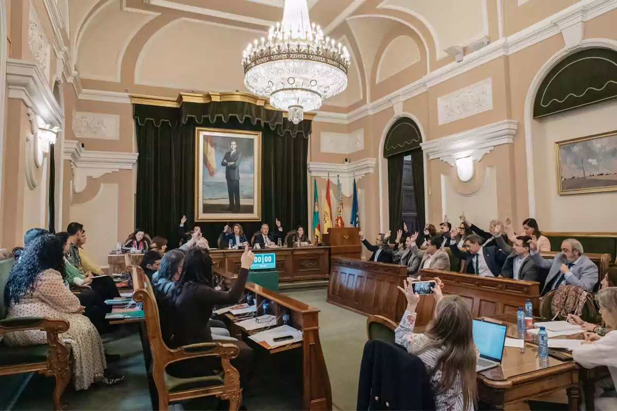 A meeting room with people sitting on wooden benches raising their hands, a large portrait on the wall, and several flags behind a main table under a chandelier.