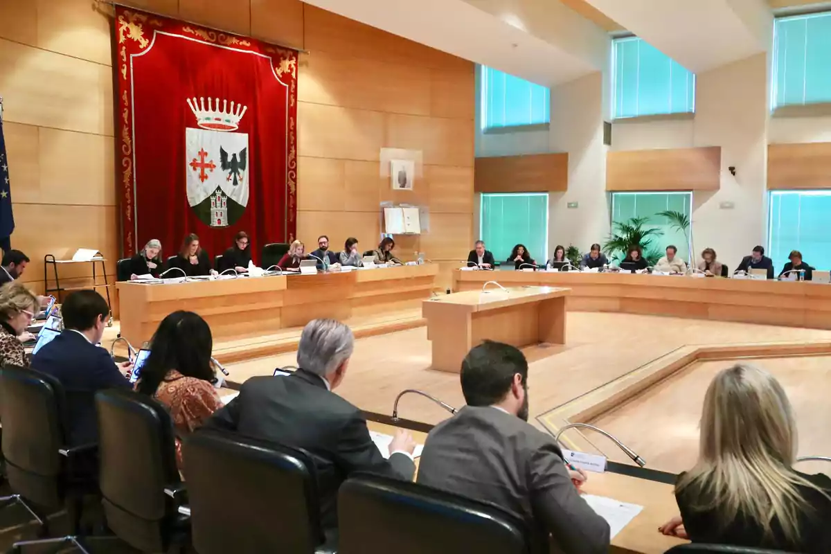 A formal meeting is held in a conference room with a large emblem on the wall and people seated around a U-shaped table.